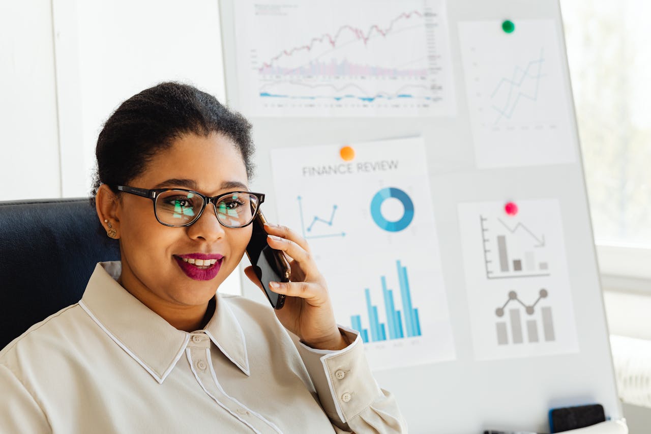 Confident businesswoman wearing glasses studies financial charts while talking on smartphone.